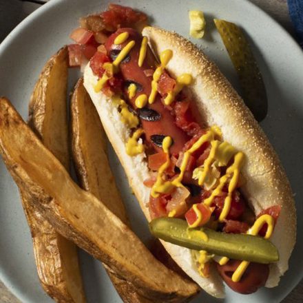 A hot dog on a toasted bun, topped with mustard, tomato, onion and sautéed onion, with a side of potato wedges.