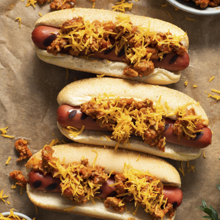 Trois hot-dogs lovés dans des pains grillés, garnis de chili et de fromage fondu, placés sur une feuille de papier parchemin et entourés d’aliments