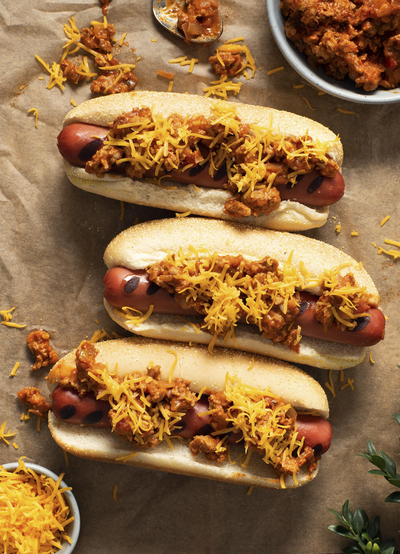 Three hot dogs in toasted buns topped with chili and melted cheese on a sheet of parchment paper, surrounded by ingredients