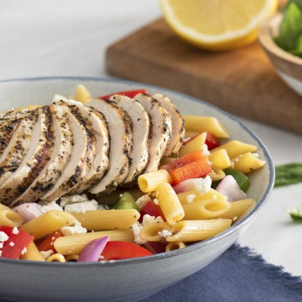 Generous strips of grilled chicken breasts on a bed of noodles, peppers, onions, and feta cheese in a bowl with a napkin, cutlery, and fresh ingredients