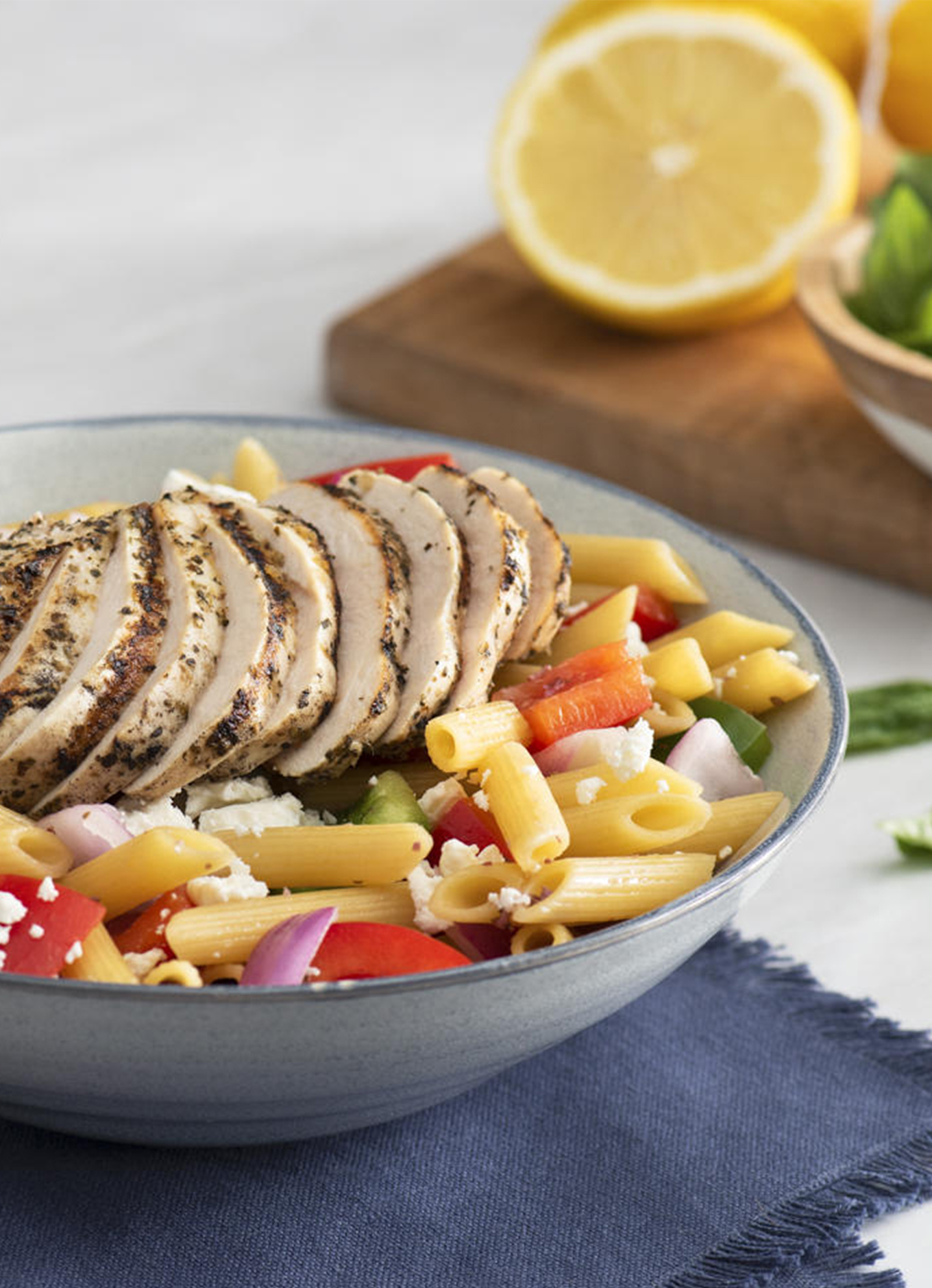Generous strips of grilled chicken breasts on a bed of noodles, peppers, onions, and feta cheese in a bowl with a napkin, cutlery, and fresh ingredients
