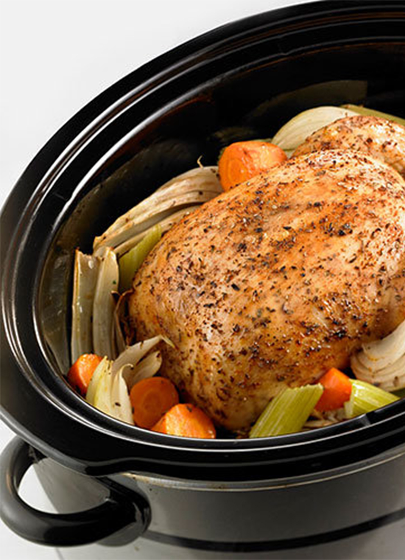 A whole cooked chicken resting in a slow cooker on a bed of celery, carrots, and onion.