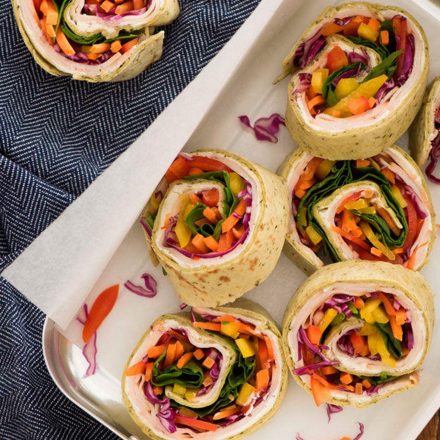 A tray of colourful Rainbow Veggie Pinwheel sandwiches, with colourful vegetables, Zabiha Halal Smoked Chicken Breast Deli slices on a wooden table.